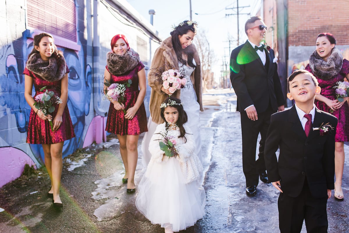 keep warm during a winter wedding | Wedding Photography | From the Hip Photo | bride in a fur coat walks down an alley. Her bridesmaids are wearing red dresses with fur scarves. Her groom and a ring boy stand by