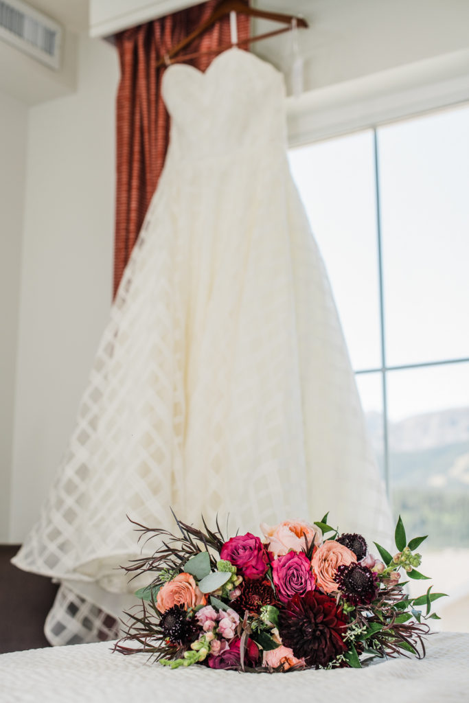 Bridal dress hangs in front of window with bouquet in the foreground