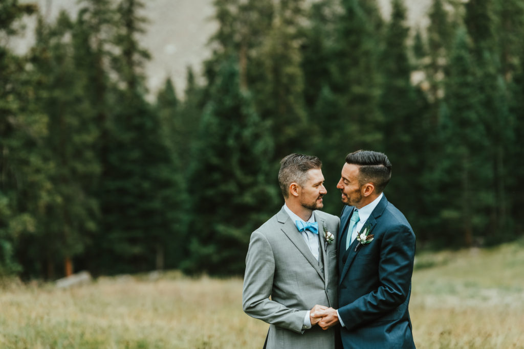 Wedding Photography | Grooms hold hands together at the edge of mountain forest