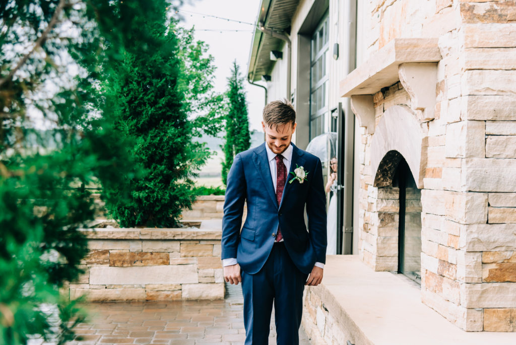 Wedding photos | Groom waits outside in the rain, with his back turned as bride appears behind him carrying umbrella