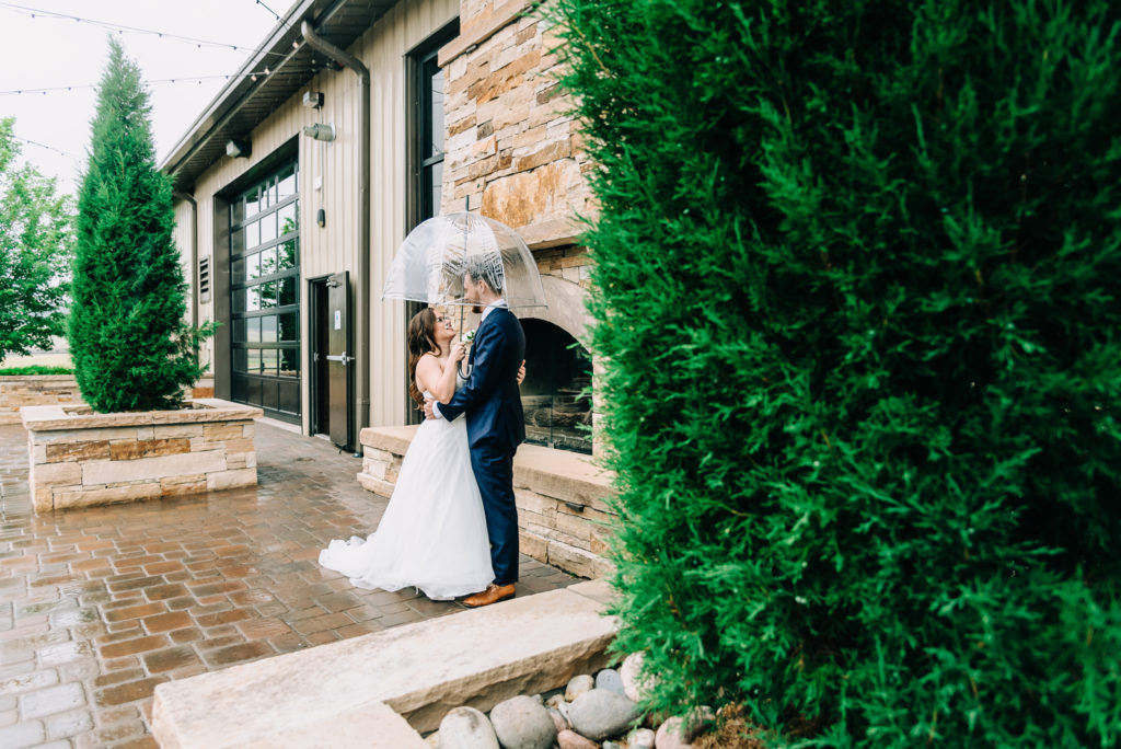 Wedding Photos | Bride holds transparent umbrella over herself and groom on rainy patio