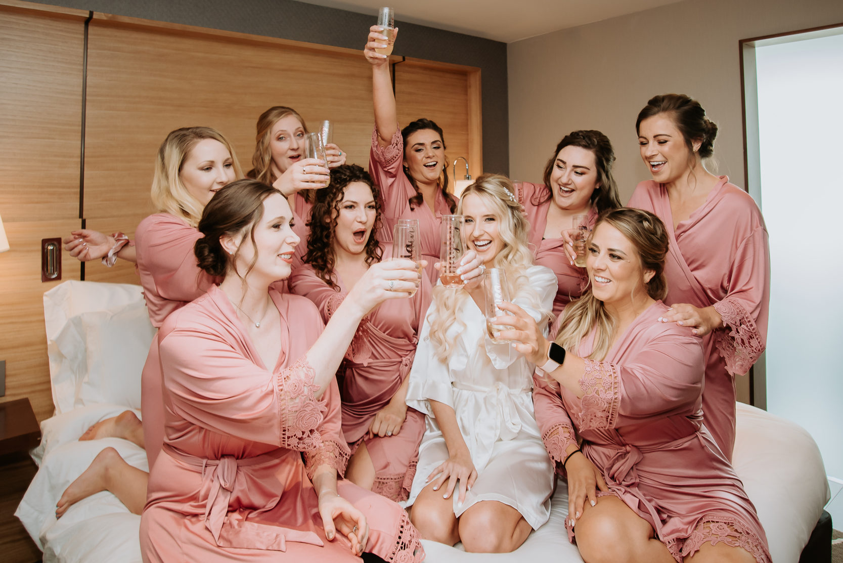 Wedding photos | Bride and bridesmaids sit on bed toasting drinks 