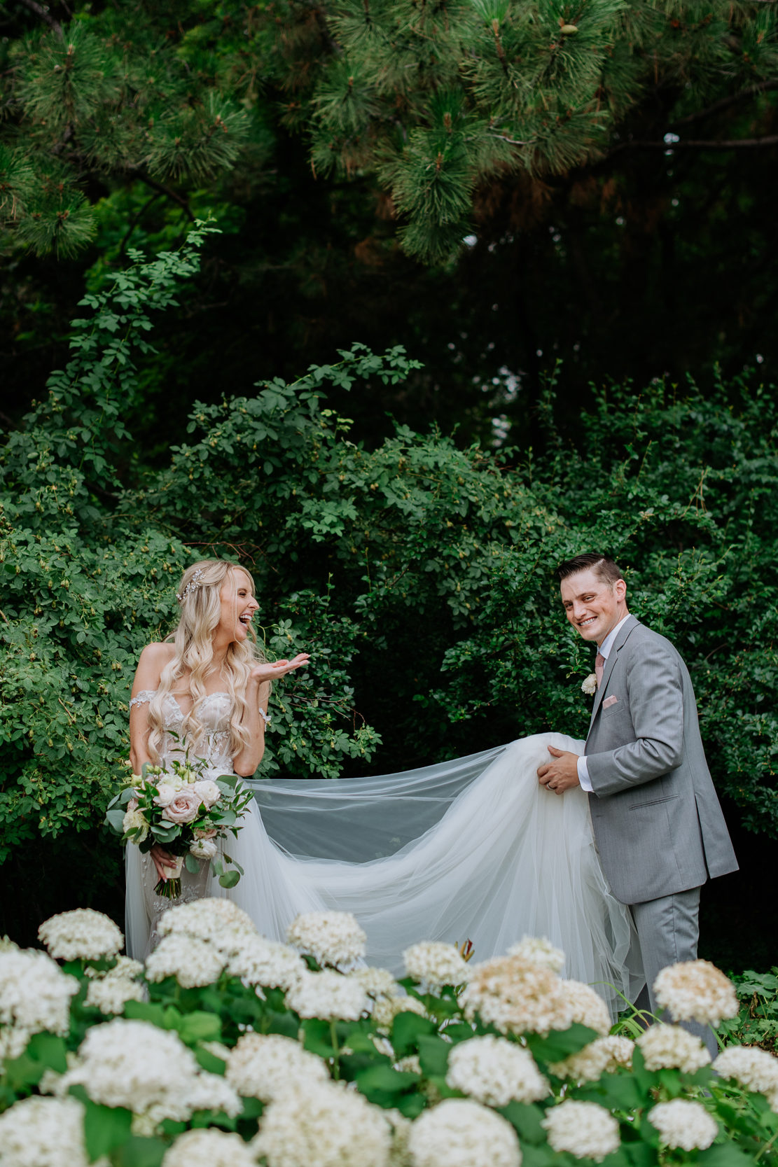 Wedding Photography | Bride blows a kiss to the smiling groom, as he carries her dress' train | Denver Botanical Gardens