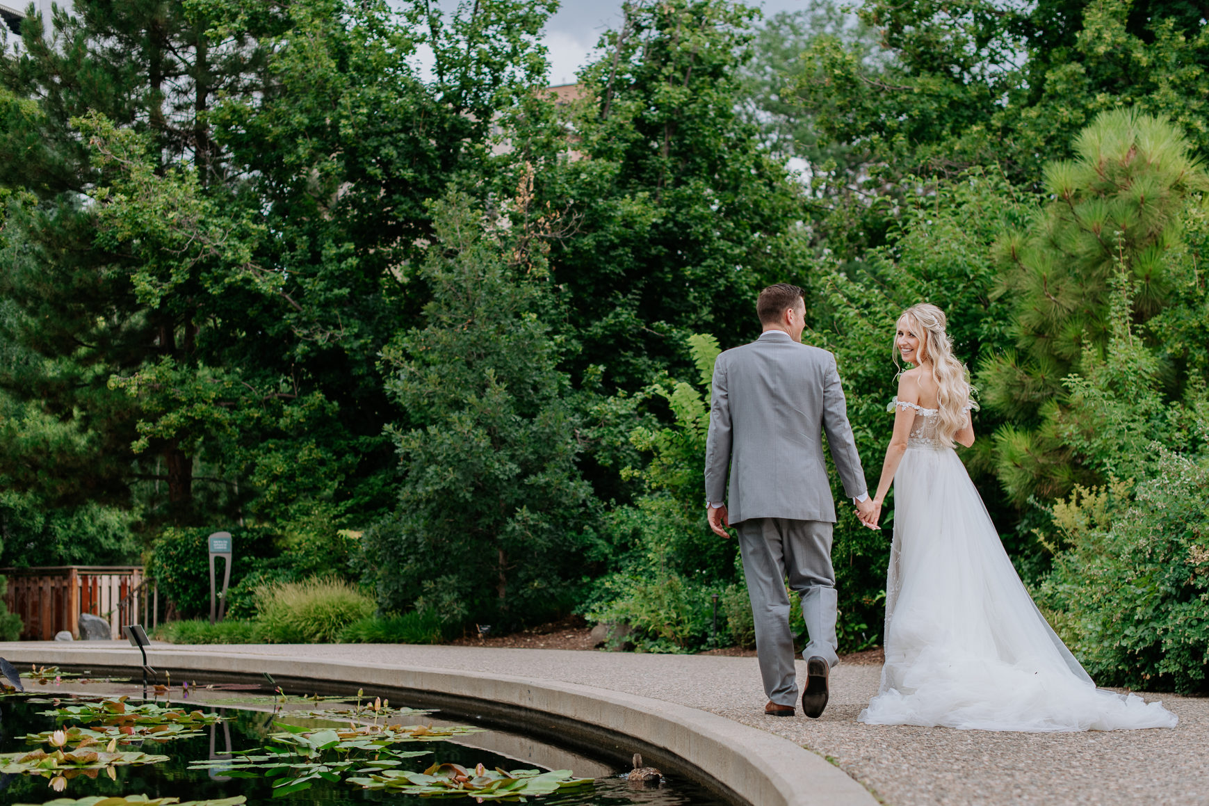 Wedding Photography | Bride glances back as she walks hand in hand with groom along lily pad pond | Denver Botanical Gardens