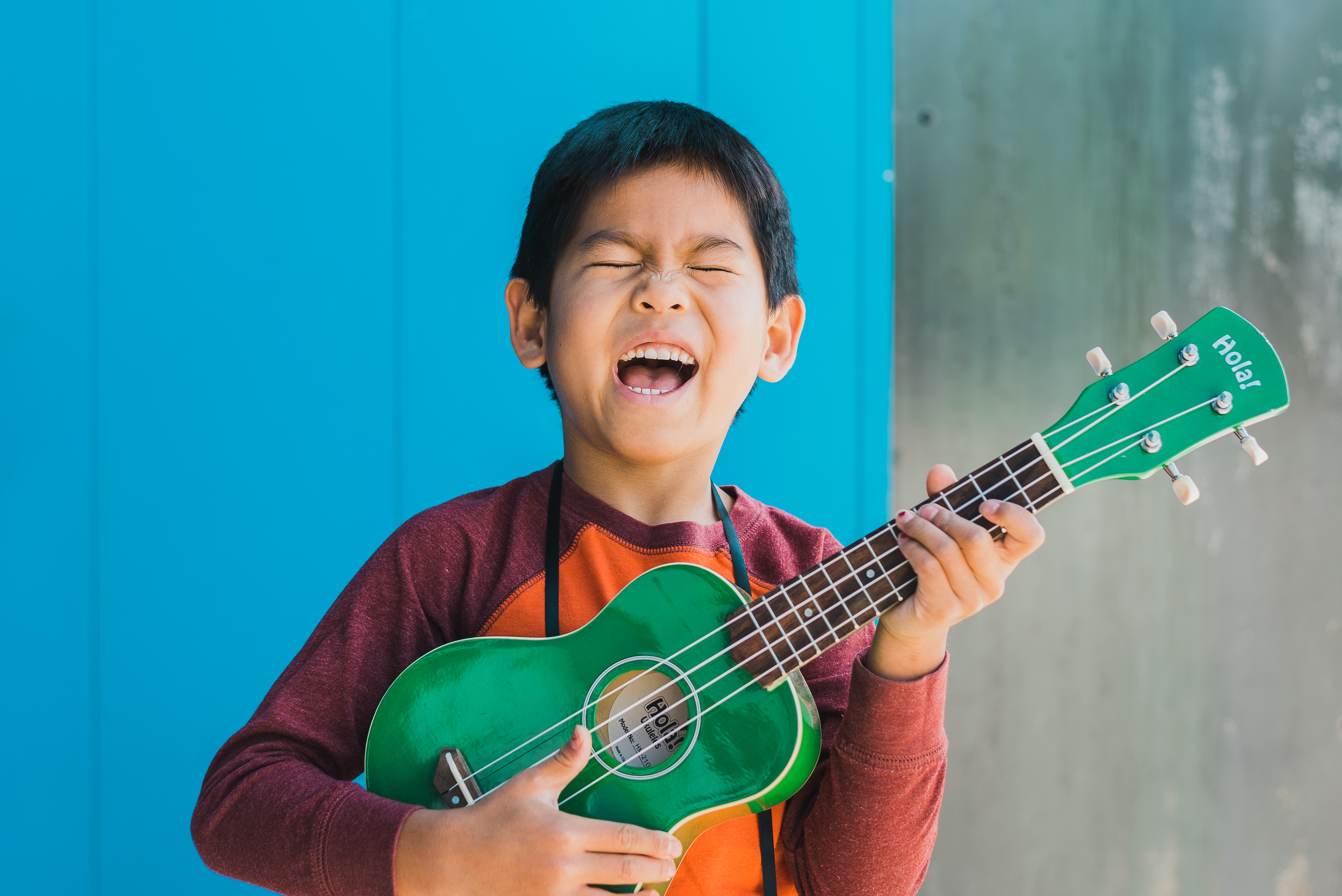 Young musician smiling with a green guitar against a blue wall at Stanley Marketplace Aurora