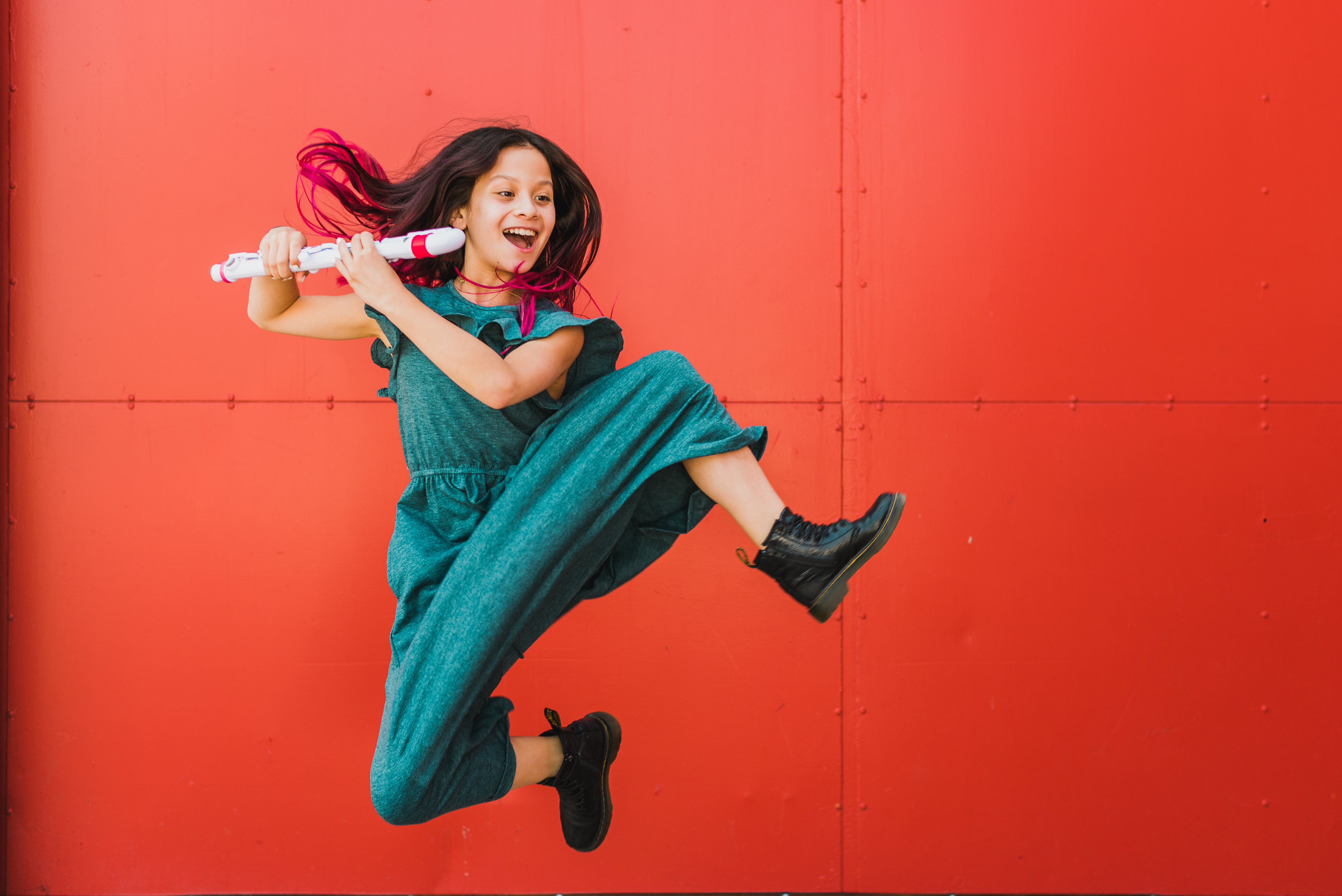 Child musician jumping energetically with a flute in front of a red wall at Stanley Marketplace